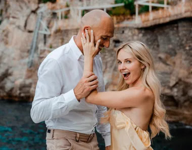 A joyful couple by the seaside, with the woman playfully touching the man's face, both smiling.