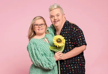 A smiling woman in a green dress and glasses holds a sunflower with a man in a colorful shirt against a pink background.