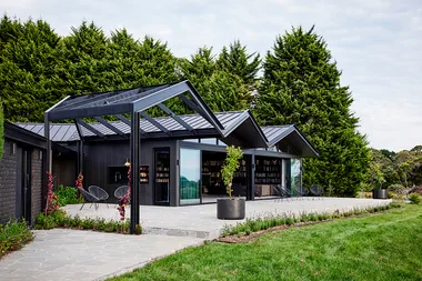 Modern black house with angular roof and large glass doors, surrounded by greenery and patio seating.