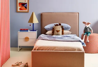 Cozy bedroom with a beige bed, lion pillow, colorful lamp, books, plush toy on a checker stool, and a framed picture.