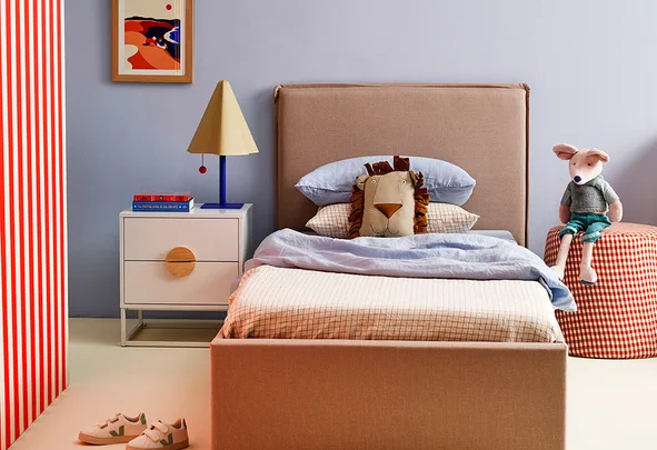 Cozy bedroom with a beige bed, lion pillow, colorful lamp, books, plush toy on a checker stool, and a framed picture.