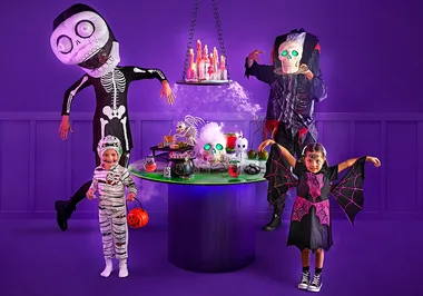 Children in Halloween costumes with skeleton and vampire, stand near a spooky table with glowing skulls and decorations.