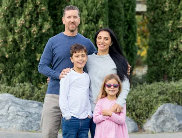 Family of four posing outdoors, green foliage in the background. The father wears a blue shirt, mother in grey, and two children.