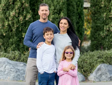 Family of four posing outdoors, green foliage in the background. The father wears a blue shirt, mother in grey, and two children.