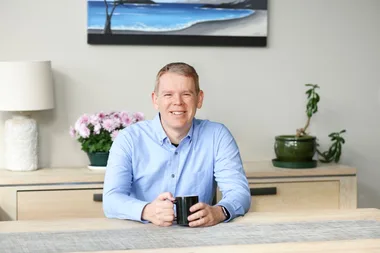 Person in a blue shirt seated at a table holding a mug, with flowers and plants in the background.