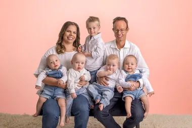 Family photo with a couple and five young children sitting on a sofa against a light pink background.