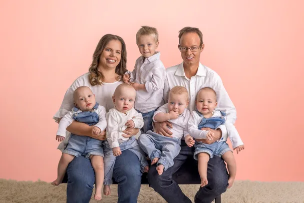 Family photo with a couple and five young children sitting on a sofa against a light pink background.