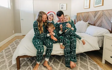 A joyful family in matching green Christmas pajamas sits on a bed, smiling and embracing each other.