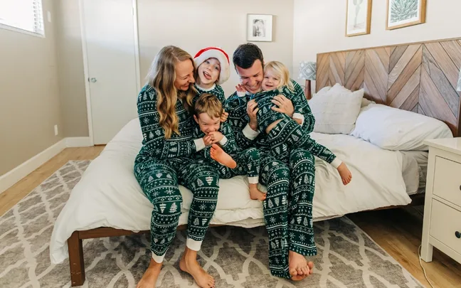 A joyful family in matching green Christmas pajamas sits on a bed, smiling and embracing each other.