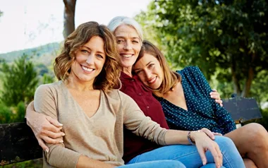 Three women seated on a park bench, smiling and relaxing outdoors with trees in the background.