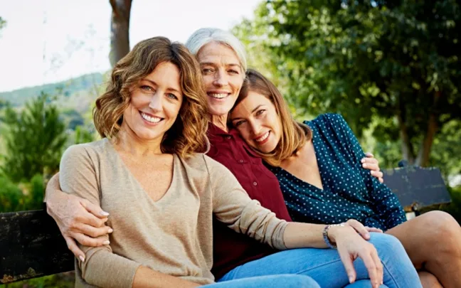Three women seated on a park bench, smiling and relaxing outdoors with trees in the background.