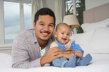 Father and baby smiling on a bed in a bright room with large windows.
