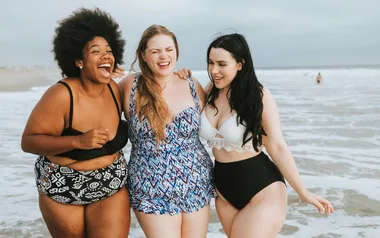 Three women in swimwear laughing and walking along the beach with the ocean in the background.
