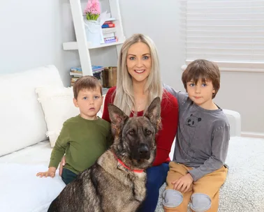 Woman with two children and a German Shepherd dog sitting on a sofa in a cozy, light-filled living room.