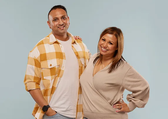 A smiling man in a plaid shirt and a woman in a beige top pose together on a light blue background.