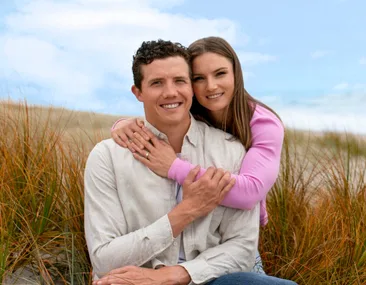 Couple sitting in a field, woman in a pink sweater hugging man in a white shirt, both smiling at the camera.