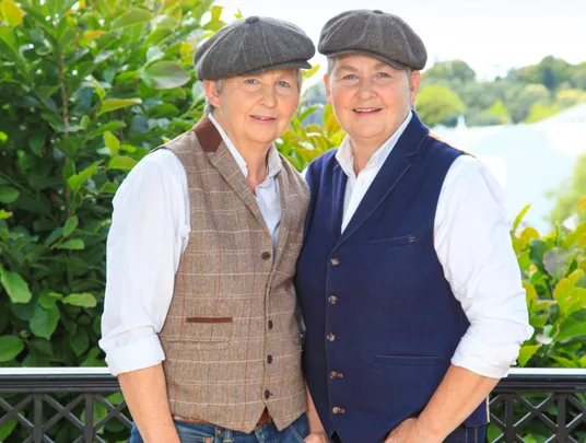 Two people wearing flat caps and waistcoats, standing outdoors in front of greenery, smiling at the camera.