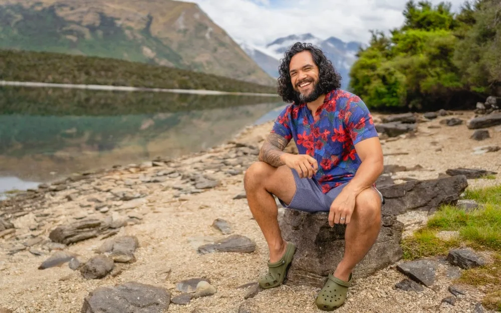 Man in floral shirt sits on rock beside a calm lake with mountains in background, smiling.