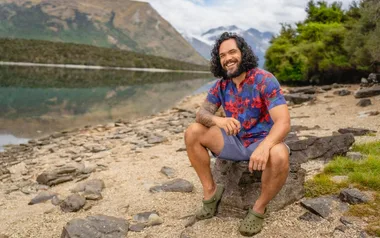 Man in floral shirt sits on rock beside a calm lake with mountains in background, smiling.