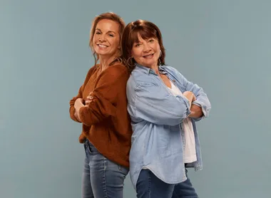 Two women standing back-to-back, smiling, with arms crossed, wearing casual shirts and jeans against a plain background.