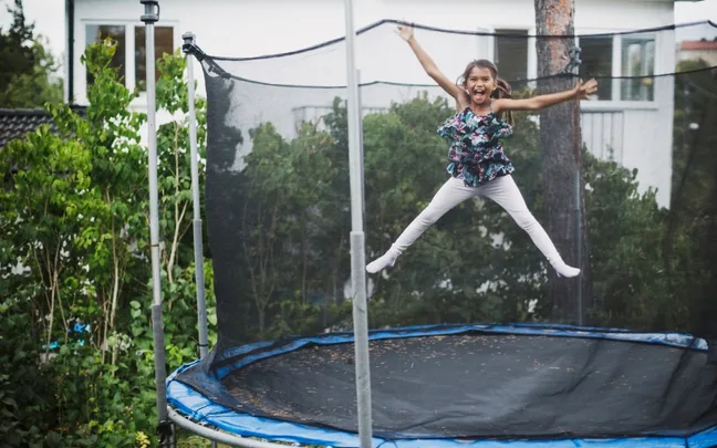 A child joyfully jumping on a trampoline in a backyard, surrounded by greenery.