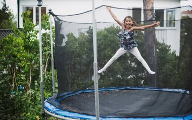 A child joyfully jumping on a trampoline in a backyard, surrounded by greenery.