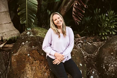 A woman in a light purple sweater sits on a rock, smiling, surrounded by lush greenery and large leaves.