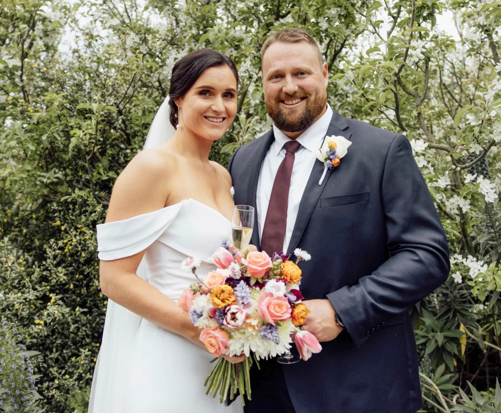 Bride and groom smiling outdoors, she holds flowers in a white dress, he wears a dark suit with a tie.