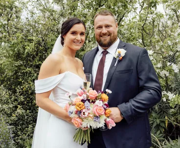 Bride and groom smiling outdoors, she holds flowers in a white dress, he wears a dark suit with a tie.