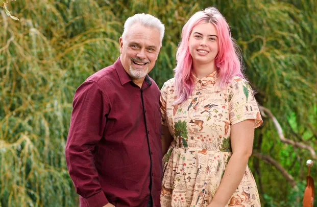 A man in a red shirt and a woman with pink hair in a patterned dress smile outdoors with a green background.