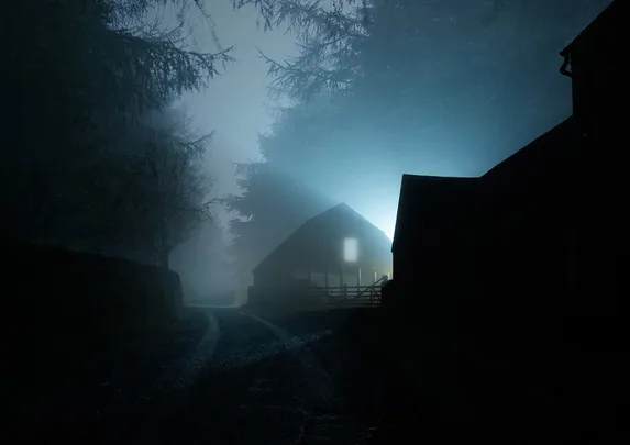 Misty night road with silhouette of a barn illuminated by blue light in the distance.