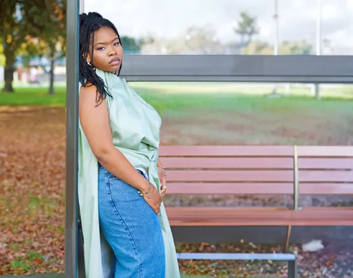 A person in a green top and jeans leans against a bus stop shelter with autumn leaves in the background.