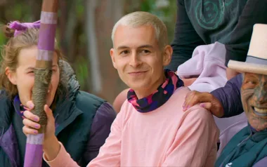A smiling person with short blond hair sits with a group, holding a colorful staff.