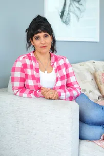 A woman in a pink checkered shirt and jeans sits on a beige sofa, with a thoughtful expression.
