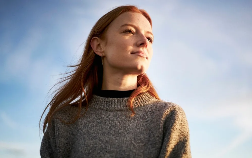 A person with long hair wearing a gray sweater looks thoughtfully into the distance under a clear sky.
