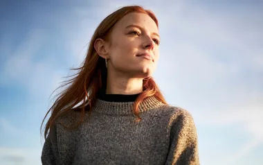 A person with long hair wearing a gray sweater looks thoughtfully into the distance under a clear sky.