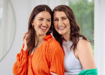 Two women smiling; one in an orange blouse, the other in a white top, standing in a bright room with a window.