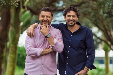 Two men smiling outdoors, one with his arm around the other, surrounded by lush greenery in a relaxed setting.