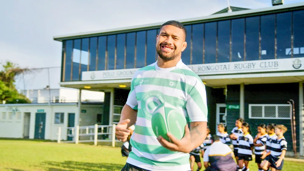 Man in striped rugby jersey holding a ball, standing in front of Rongotai Rugby Club building. Children in background.