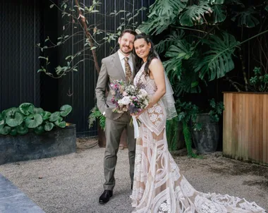 Bride in intricate lace gown and groom in a suit, standing together with a bouquet, in a lush garden setting.