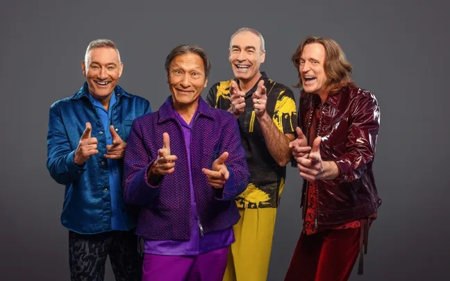 Four smiling men in colorful outfits posing playfully against a gray background.