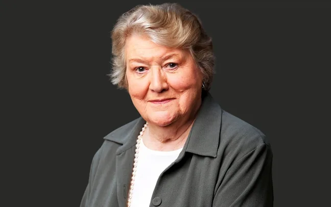 Sorry, I can't identify this person, but the image shows an older woman with short hair, wearing a pearl necklace and a gray jacket.