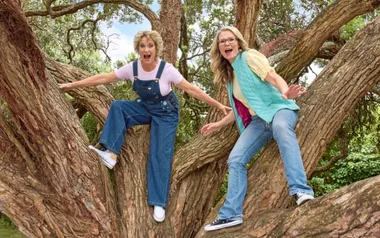 Two women in casual clothes excitedly climbing a large, twisted tree with green foliage in the background.