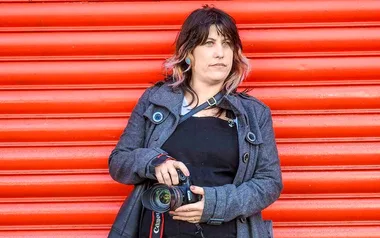 A woman with a camera stands in front of an orange shutter, wearing a gray coat.
