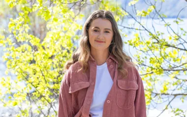 A person with long hair smiles outdoors, wearing a pink jacket and white top, surrounded by leafy branches and sunlight.