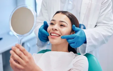 A woman smiles at a dentist's office, holding a mirror, assisted by a dentist wearing blue gloves.