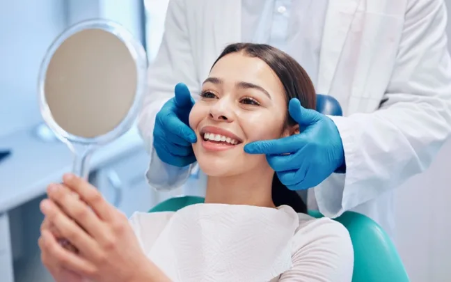 A woman smiles at a dentist's office, holding a mirror, assisted by a dentist wearing blue gloves.