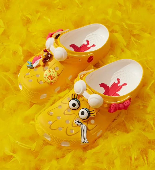 Yellow crocs with cartoon eyes and decorative charms, including a character and a bow, on a fluffy yellow background.