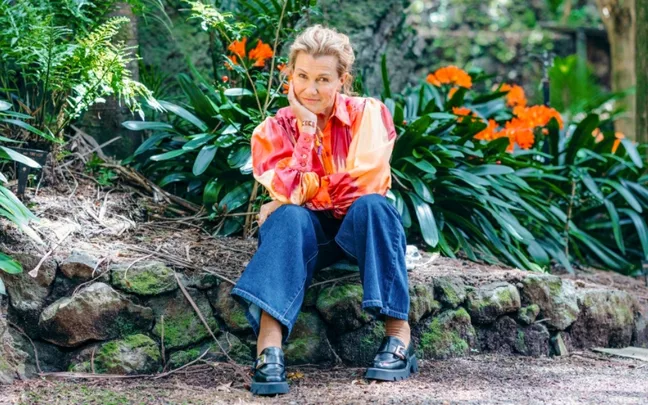 Woman in colorful blouse and jeans sits on stone ledge in garden, surrounded by green plants and orange flowers.