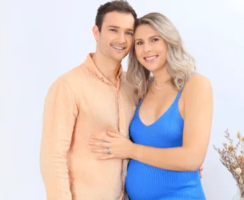 Couple smiling, woman in blue dress with visible pregnancy, man in peach shirt, white background with dry flowers.
